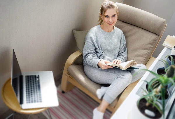 Junge Frau mit Buch in der Hand sitzt auf beigem Sessel in Wohnung — Stockfoto