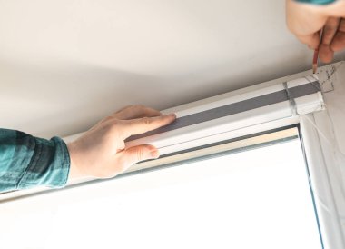 Man installs gray blackout roller blinds on window indoors, closeup.