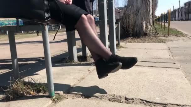 Девушка сидит на скамейке на автобусной остановке и трясет ногами в предвкушении — стоковое видео