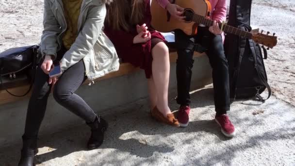 Varias chicas y un tipo que toca la guitarra, se sientan en el banco y cantan canciones. Jóvenes al aire libre — Vídeos de Stock