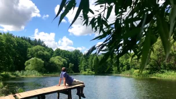 Relaks na całe życie, koncepcja naturalnej terapii. Młodzi ludzie siedzący relaksuje się do huśtawka stóp w pobliżu powierzchni wody na krawędzi bocznej drewnianej w przyrodzie rzeka jezioro w pięknym letnim dniu, Slow Motion shot — Wideo stockowe