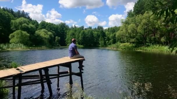 Отдых на всю жизнь, концепция природной терапии. Молодой человек сидит расслабляет качели возле поверхности воды на боковом краю деревянного на озере реки природы в красивый летний день, замедленной съемки — стоковое видео