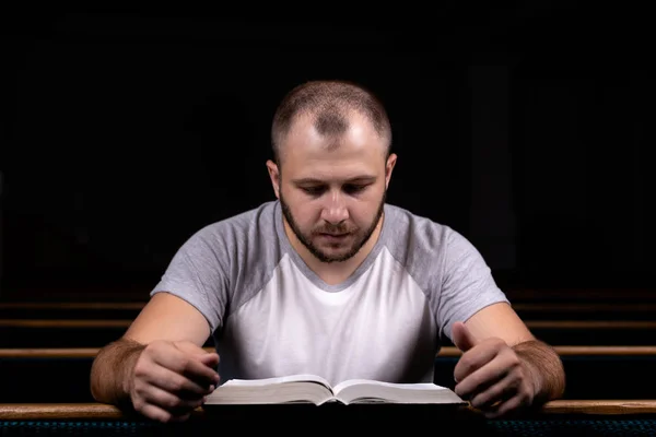 A young guy sits on a church bench, reads the bible and prays. T