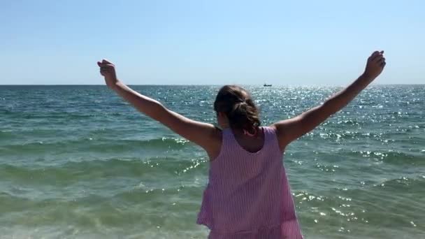 Κλείστε την πλευρική όψη μιας νεαρής καυκάσιας που στέκεται σε μια παραλία με τα χέρια του στον αέρα, κοιτάζοντας έξω στη θάλασσα — Αρχείο Βίντεο