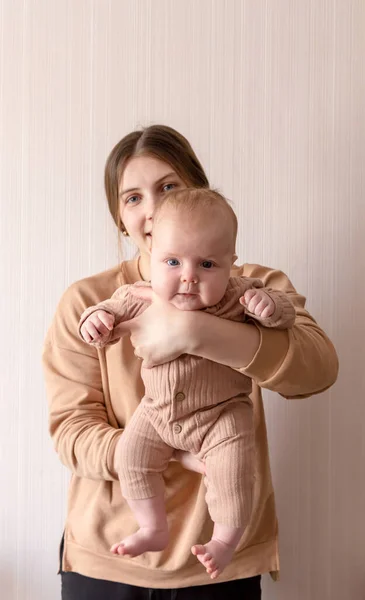 Молодая мать держит на руках девочку, которой 3 месяца. Мама и ребенок в коричневой одежде на светлом фоне — стоковое фото