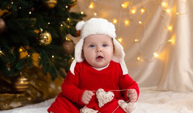 Noel bebeği kameraya bakıyor. Kırmızı elbiseli ve beyaz şapkalı şirin küçük bir kız duygularını ifade ediyor. Arka planda küçük çocuk, ağaç ve çelenk olan Noel konsepti bulanık