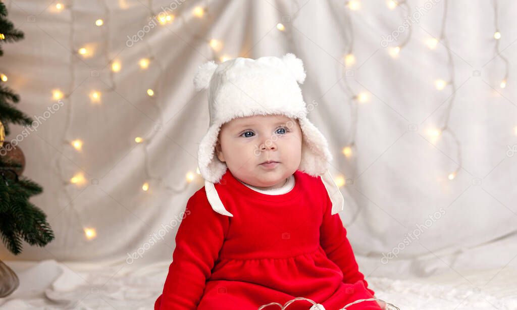 Christmas child is thoughtful, dreamily surprised emotions. A cute little girl in a red dress and white hat expresses emotions. Christmas concept with little kid tree and garland on background in blur