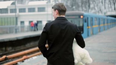 Genç adam elinde bir oyuncak ayı tutuyor. Giden metro vagonuyla ilgileniyor..