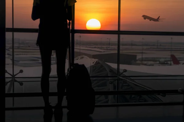 Silhouette of young woman is standing near window at the airport and watching plane before departure. She is looking at airplane taking off and carrying luggage.Travel Concept .