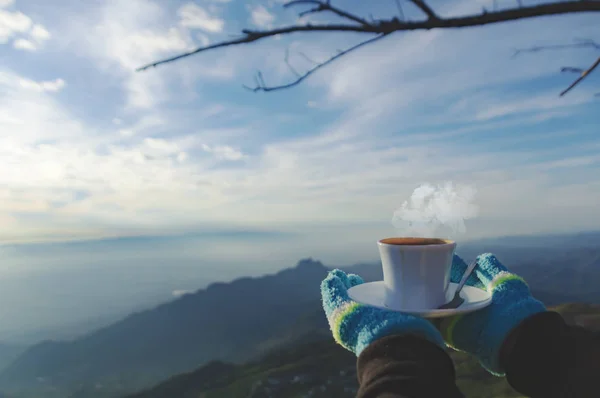 Closeup a cup of coffee in traveler\'s hand over out of focus mountains view. A young tourist woman drinks a hot drink from a cup and enjoys the scenery in the mountains.  - Image