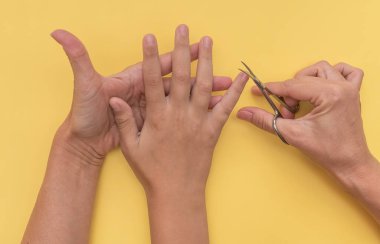Yetişkin beyaz kadın sarı arka planda çocuğunun tırnaklarını kesiyor.