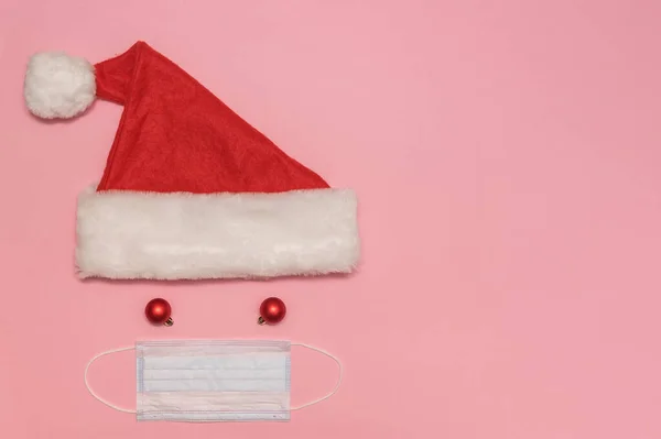 Kırmızı Noel Baba şapkası, ponpon ve maskeli. Oyuncak toplar ve koronavirüs için maske. Pembe bir arka planda.