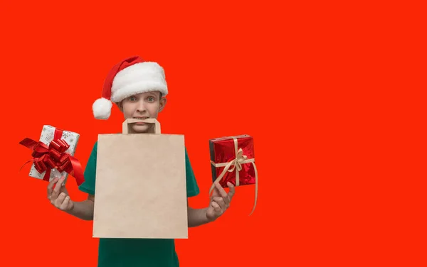 一个穿着绿色T恤的孩子 一双惊讶的眼睛盯着红色背景 手里拿着礼物 带着手提包和圣诞帽的男孩 — 图库照片