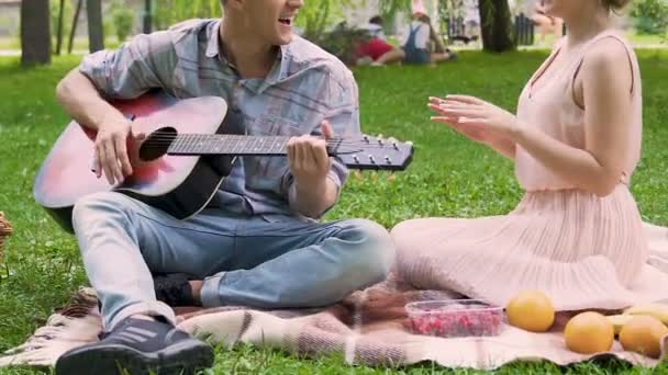 Muž hraje kytaru a zpívat píseň s přítelkyní parku piknik, zbytek přátel — Stock video