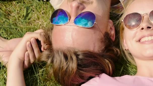 Šťastný pár ležet na trávě v sluneční brýle a drželi se za ruce, obloha reflexe — Stock video
