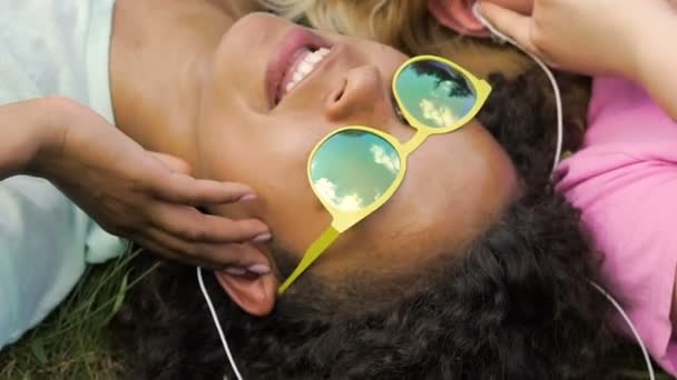 Amigas felizes em fones de ouvido ouvindo música juntas, deitadas na grama — Vídeo de Stock