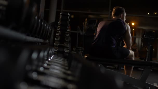 Atleta masculino cansado após o treino senta-se no banco, palpita-se sozinho no ginásio à noite — Vídeo de Stock