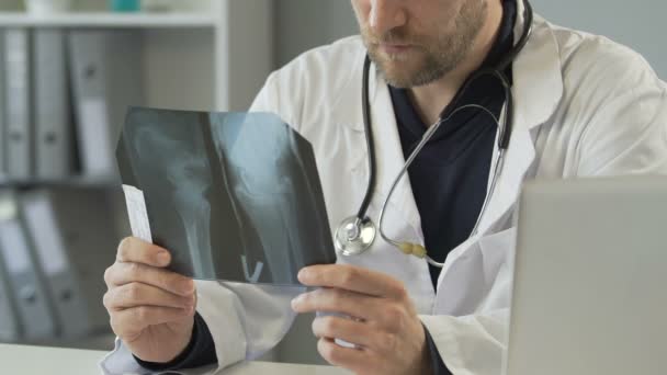 Ortopedista experiente olhando para raio-x perna e paciente de consultoria no laptop — Vídeo de Stock