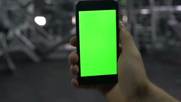 Чоловіча рука тримає смартфон у тренажерному залі, фітнес-додаток для бодібілдингу, зелений екран — стокове відео