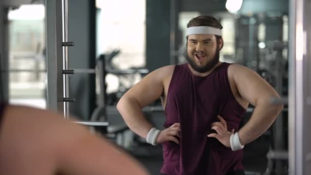 Смешной толстяк, смотрящий на зеркальное отражение спортзала и позирующий, притворяющийся мускулистым — стоковое видео