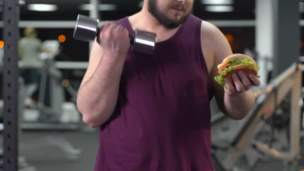 Gruby mężczyzna podnoszenia hantle i patrząc na hamburger z drugiej strony, siły woli — Wideo stockowe
