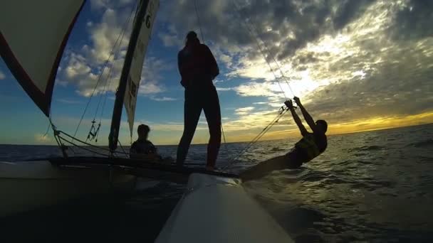 Silhouetten von drei Personen, die auf einem Windsurf-Katamaran gegen den Sonnenuntergang segeln — Stockvideo