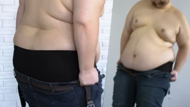 Толстый мужчина пытается надеть узкие джинсы, проблемы с избыточным весом, нездоровый образ жизни — стоковое видео
