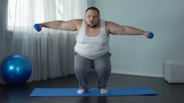 Uomo sovrappeso accovacciato e sollevamento manubri su tappetino, casa di allenamento corpo pieno — Video Stock