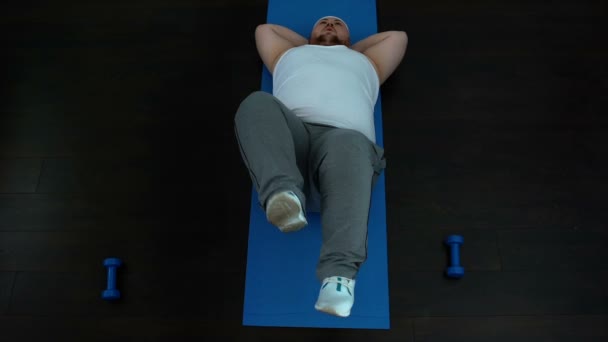 Немотивированный жирный мужчина лежит на коврике делая упражнения на велосипеде, проблема потери веса — стоковое видео