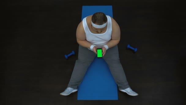 Homme joufflu utilisant l'application de remise en forme sur smartphone, assis sur le tapis, écran vert — Video