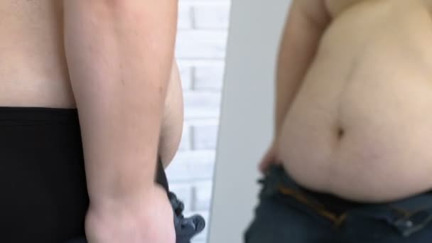 Feta ung man som försöker sätta på jeans i butiksinredning rum, stor storlek, diet — Stockvideo