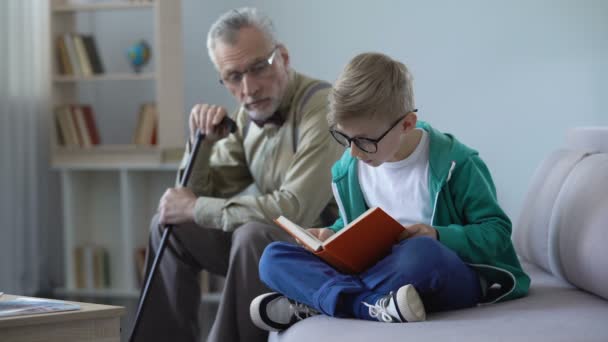 Niño en gafas libro de lectura para el abuelo, hombre viejo que se queda dormido en el sofá acogedor — Vídeo de stock