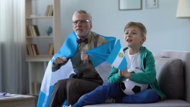 Opa zwaaien Argentinië vlaggen samen met kleinzoon, voetbal thuis kijken — Stockvideo