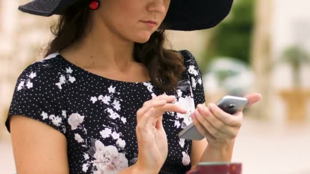 Gyönyörű hölgy kalapban ül utcai kávézó, görgetés okostelefon-alkalmazás — Stock videók