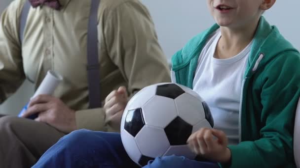 Ребенок с дедушкой вместе смотрят футбольный матч и поддерживают любимую команду — стоковое видео