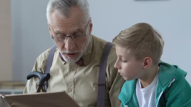 Мужчина-пенсионер читает сказки для внука, объясняет школьную тему, помогает — стоковое видео