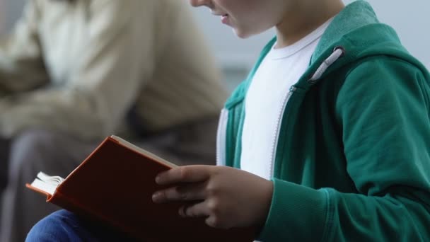 Niño sosteniendo libro aprendiendo a leer con el padre sentado en el sofá, lección con tutor — Vídeos de Stock