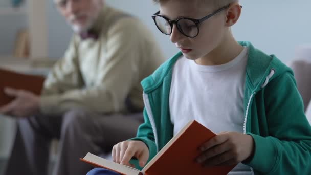 Gracioso niño pequeño en gafas libro de lectura con abuelo feliz detrás, ratón de biblioteca — Vídeo de stock