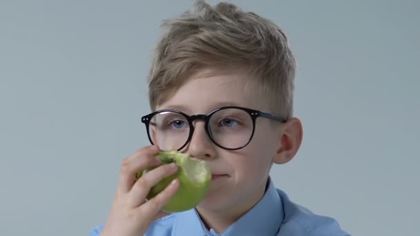 Смешной мальчик в очках, кусающий зеленое яблоко, здоровое детское питание, витаминные закуски — стоковое видео
