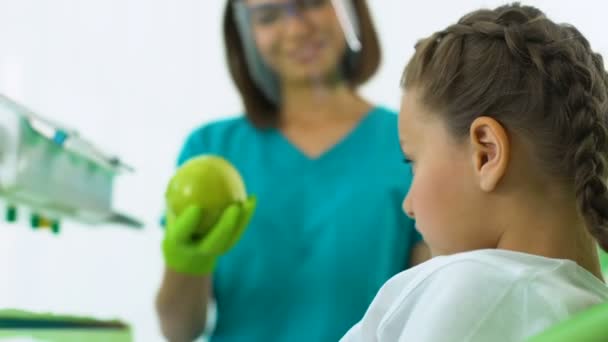 Crianças dentista dando maçã para menina, pequeno paciente comendo frutas com prazer — Vídeo de Stock