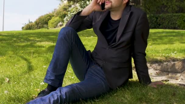 Бизнесмен разговаривает по телефону в парке, сидит на газоне, отдыхает после рабочего дня — стоковое видео