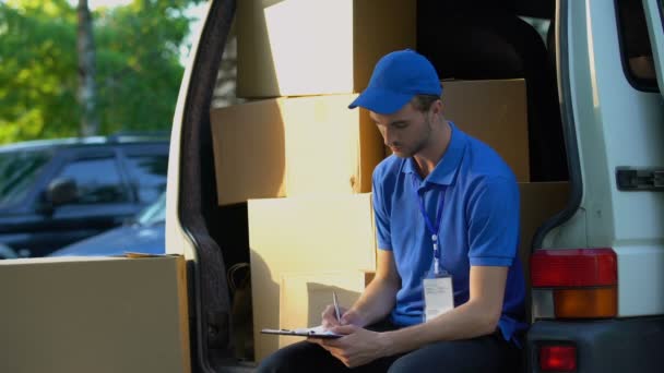 Νέοι deliveryman γράφοντας αγροτεμάχιο αναφορά κατανομής και χαμογελαστός, κάθεται σε φορτηγό — Αρχείο Βίντεο