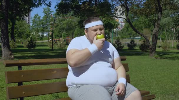 Толстый молодой человек ест яблоко после пробежки на свежем воздухе, надлежащий обед с витаминами — стоковое видео