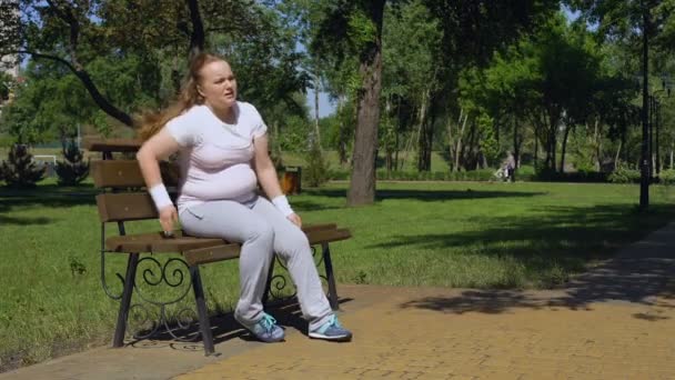 Толстая девушка сидит на скамейке, уставшая после пробежки, но продолжает бежать, мотивация — стоковое видео