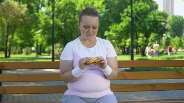 Dik meisje worstelt met de verleiding om te eten van de hamburger, liever junkfood, geen wilskracht — Stockvideo