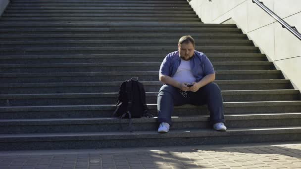 Товстий чоловік слухає музику на сходах, самотність, надмірна вага викликає невпевненість — стокове відео