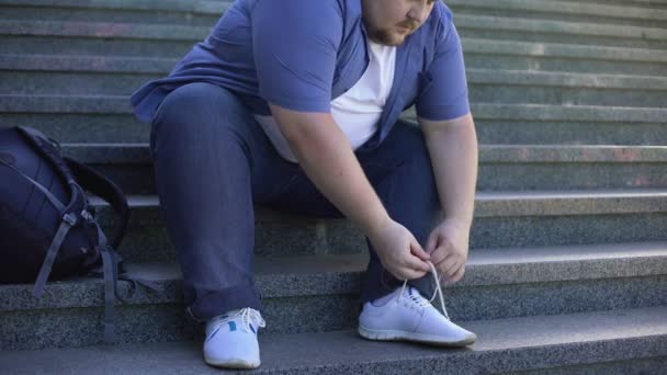 Moeilijk voor dikke jonge man te binden schoenveters, uitdagingen zwaarlijvige mensen gezicht elke dag — Stockvideo