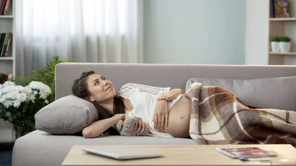 Expecting woman lying on couch with teddy bear, thinking about future child