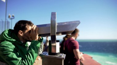 Young man looking through binoculars at observation deck, admiring sea horizon clipart