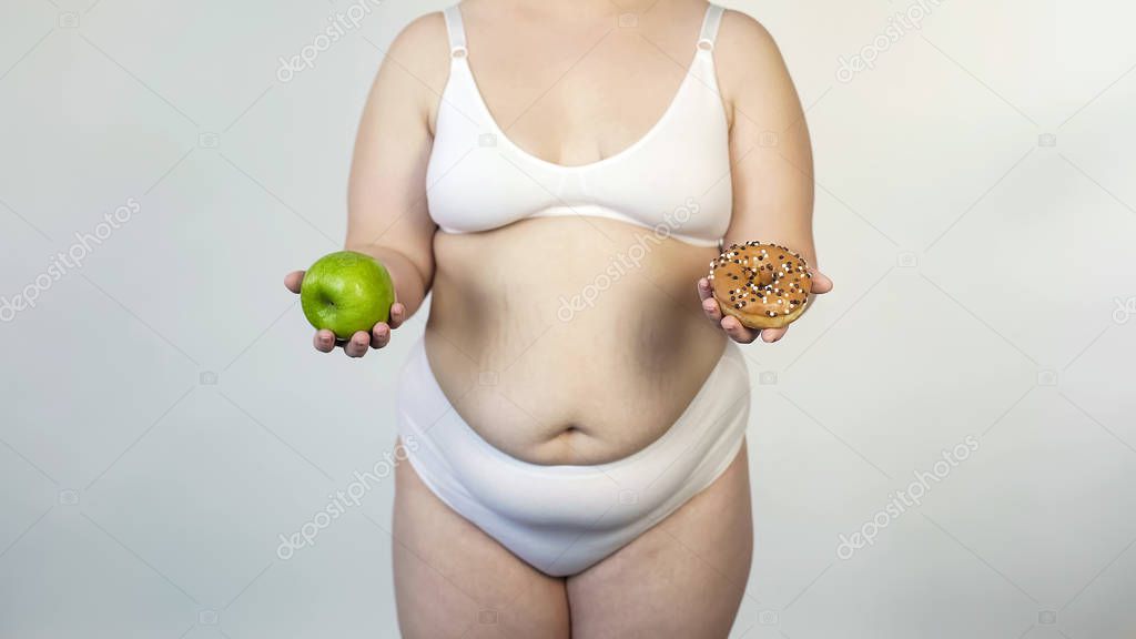 Corpulent woman showing apple and donut, nutrition choice, diet or overeating